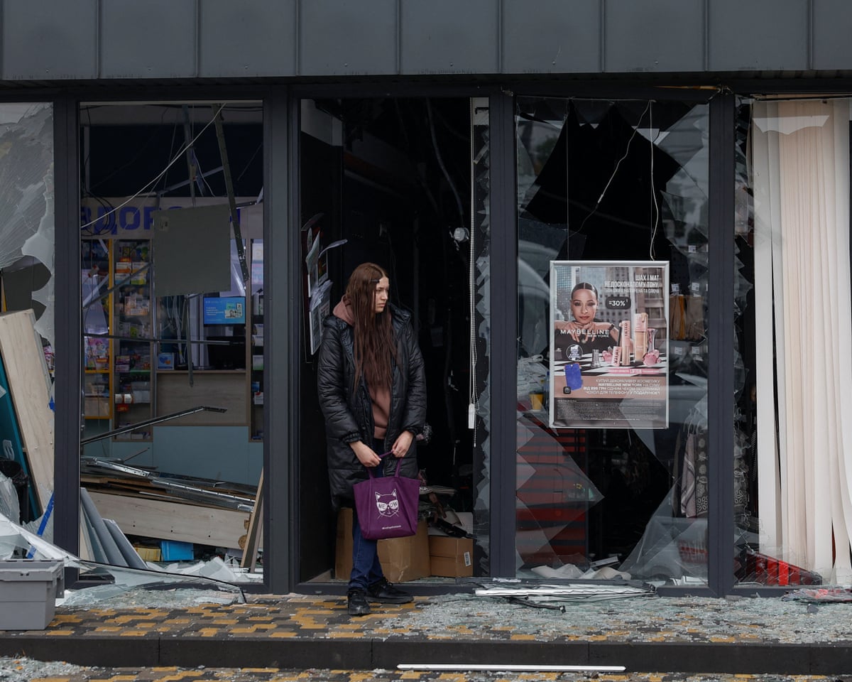 A woman holding a bag stands amid shattered glass outside a missile-damaged Kyiv storefront