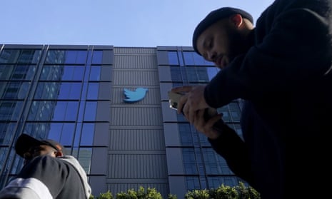 Two men walk outside a building that bears the Twitter blue bird logo. One man is staring at his mobile phone.