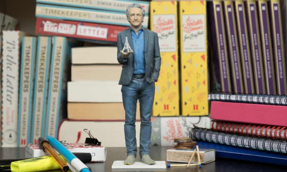 A miniature model of Simon Garfield standing on a desk holding a miniature of the Eiffel Tower, normal size books behind him