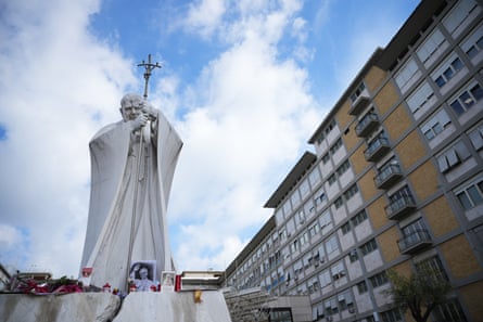 A marble statue of St John Paul II with candles and photos at its base outside a hospital