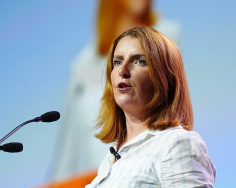 A woman with shoulder-length hair and wearing a shirt speaks in front of a microphone.