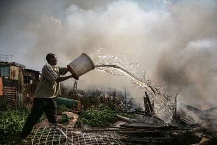 A man throws water from a bucket onto burning remains of a shack.