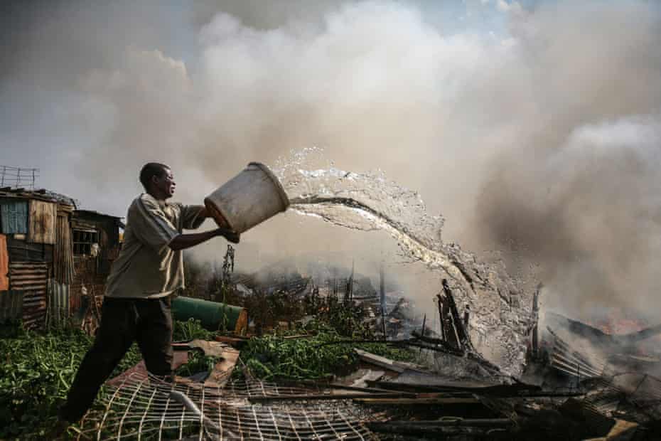 A man pours water from a bucket onto the burning remains of a hut.