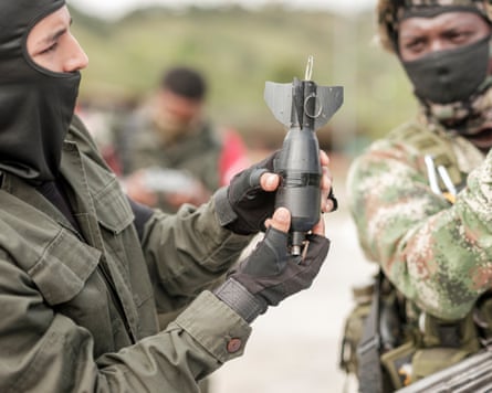A person in military clothing holds up a grenade to show another person. Both wear balaclavas.