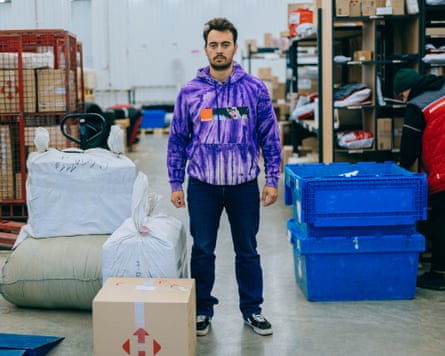 A man with dark hair and wearing a purple jumper stands in a warehouse next to large bales and boxes