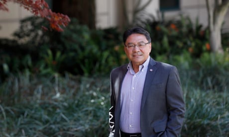 An Asian man with black hair and glasses, smiling and looking casual with his hands in his pockets, in a blue suit standing outside a building with lots of greenery.