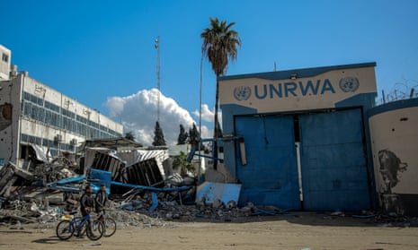 two people wheel bikes past rubble and a building painted in blue and white with sign UNRWA