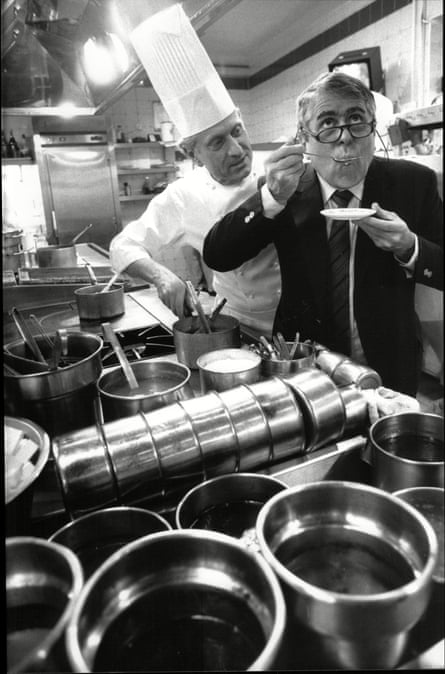 Michel Roux, dressed in chef’s whites and chef’s hat, looks on as his brother Albert, dressed in a suit, tastes his cooking in a professional kitchen