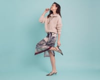 A woman in a white quarter-zip jumper, cream and paisley wrap skirt, pendant necklace and heels against a blue background.