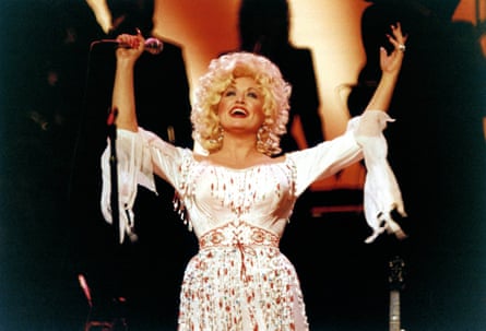 Dolly Parton, with curly blonde hair in a beaded white dress, raises her arms on stage