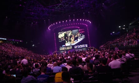 A venue filled with people. In the distance is a stage lit up in purple with a large video screen showing a team of gamers sitting at a table