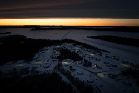 A truck traverses a frozen lake leading to the small community, which is seen in the near-darkness with a few buildings illuminated. The sun is rising on the horizon behind the lake in the background.