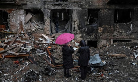 Two people dressed in black, one holding a pink umbrella, inspect the charred ruins of their home