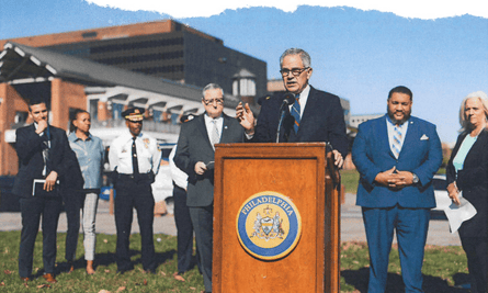 A man wearing glasses, a suit and tie, makes an address outside as people listen