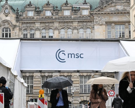 People with umbrellas walk past a pedestrian bridge with the logo of the Munich Security Conference leading to the hotel "Bayrischer Hof" in Munich, Germany.