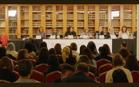 People sit at a long table on a platform as an audience looks on