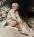 A young blond woman in a cream coloured cardigan sitting on a towel on a beach in front of the entrance to a cave