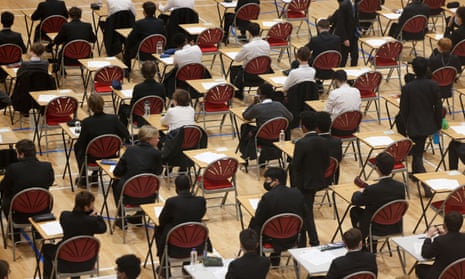 Angled view from above of rows and rows of desks in a large exam hall with boys sitting at them, some evidently wearing face masks