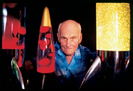 Edward Craven Walker poses between lava lamp of different colours