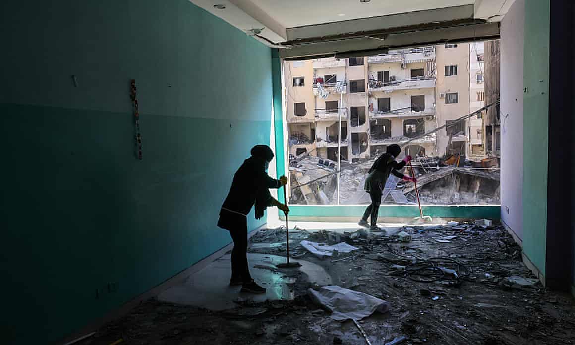 People clear debris from a room, which has no windows and faces a damaged building