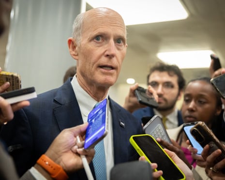 A thin bald man speaks into smartphones among a gaggle of people.