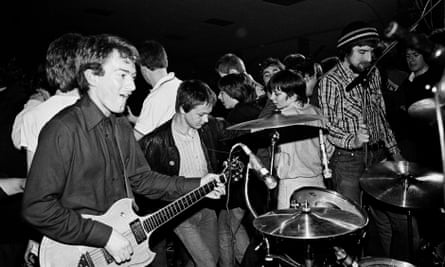 Black and white photo of a young Andy Gill playing guitar near the drum kit, very close to dancing members of the audience behind him
