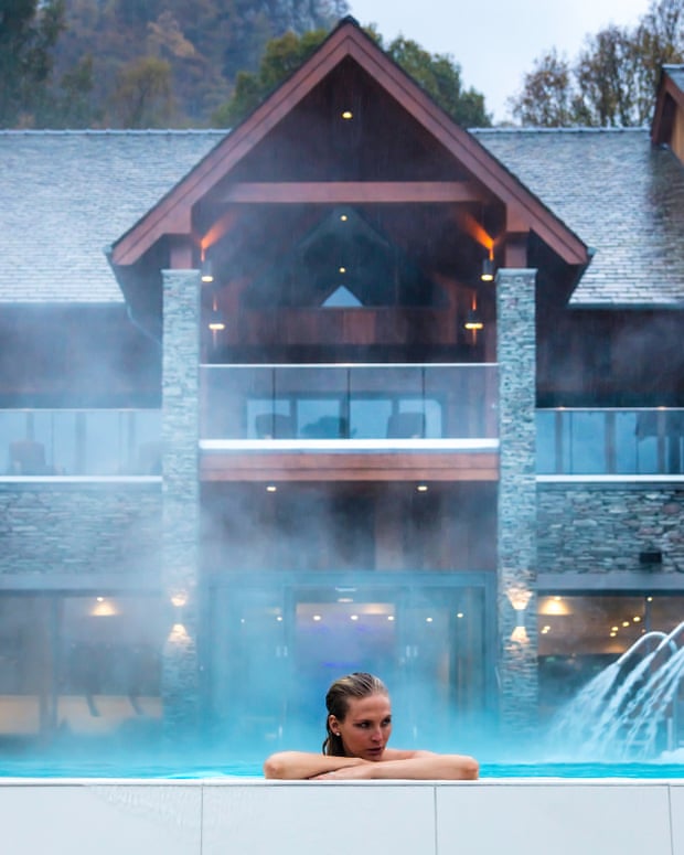 A woman rests on the edge of an outdoor pool while steam rises, with a hotel and trees in the background