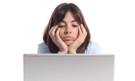 A bored young woman sits in front of her laptop screen with her face resting on her hands