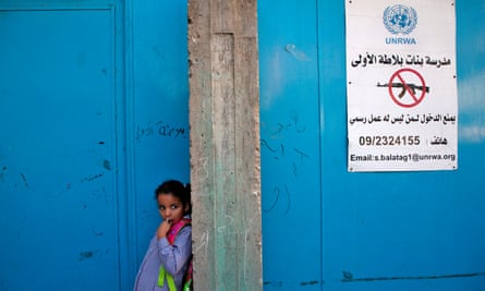 A girl stands at the entrance to a school - two large blue doors, next to a sign with the UN logo that forbids firearms