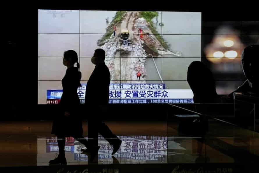 People wearing face masks walk by a TV screen at a shopping mall in Beijing showing footage of rescuers fortifying a temporary dyke built to hold back floodwaters in China’s Shanxi province
