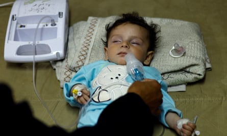 A toddler lies on the floor with a blanket under their head and is being fed through a drip. They have their eyes closed and are wearing a light blue top.