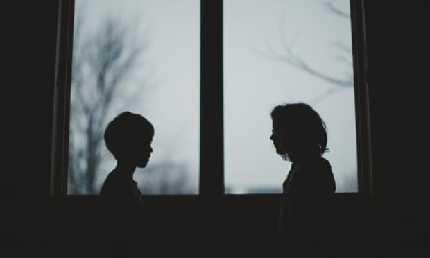 Little boy and little girl stand in front of a window looking at each other creating two silhouettes in of the window with overcast skies behind them.