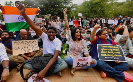 People sitting on the ground chanting, holding banners and an Indian flag