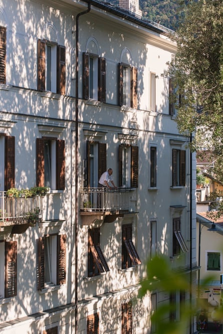 An elderly man stands on the balcony of a residential building in Bolzano, in the shadow of trees that protect him from the direct summer sun. Italy, 2022