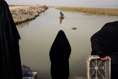 Three women in black robes are seen from the back, waiting in front of marsh area where there is a small fishing boat