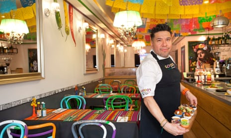 Rafael Onate in dark blue chef's apron, carrying a board of dips in colourful dining room at Gordo's