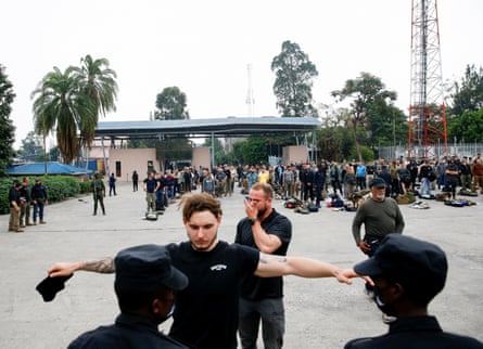 Two guards perform a security check on a man wearing a black T-shirt standing with his arms held out to the sides. Long lines of other men queue behind them