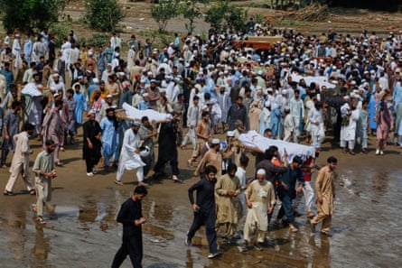 A crowd of people walks along muddy ground, with mourners carrying bodies wrapped in white sheets on stretchers on their shoulders