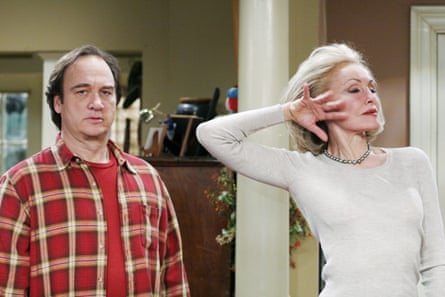 A blond woman hold her hand to her cheek with her eyes closed as a man in a red checked shirt stands next to her.