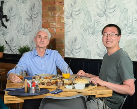 A man with white hair and a blue shirt and a younger man with dark hair and glasses and a green T-shirt sitting eating in a restaurant