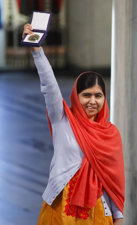 Malala Yousafzai wearing an orange dress, pale-purple cardigan and red head scarf, smiling and holding up her Nobel medal