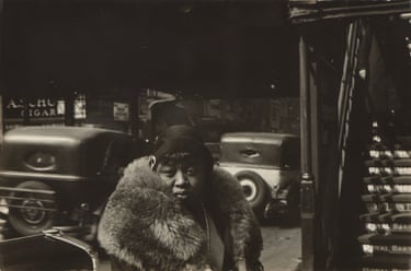 A woman in a dramatically fur collared coat pictured next to a subway entrance on a city street at night