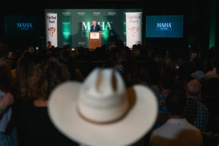 A large cowboy hat blurred in the foreground, with a man at a lectern in front of a green curtain fair in the background across a darkened hall.