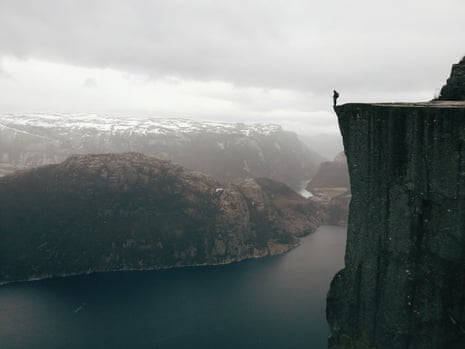 A man standing right on the edge of a steep cliff, with snowy mountains in the background, in Norway