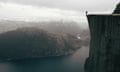 A man standing right on the edge of a steep cliff, with snowy mountains in the background, in Norway