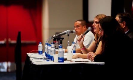 Seen from the side, a long table with a water bottle for each person, and a woman in the middle speaking into a microphone.