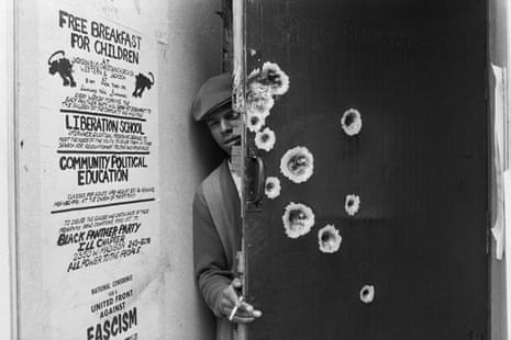 Black Panther peers from behind a door that has at least 10 large bullets holes. On the wall is a poster advertising Black Panther programs.