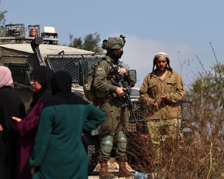 Palestinian women on left blocked from their olive groves, with Israeli soldier and settler looking on.