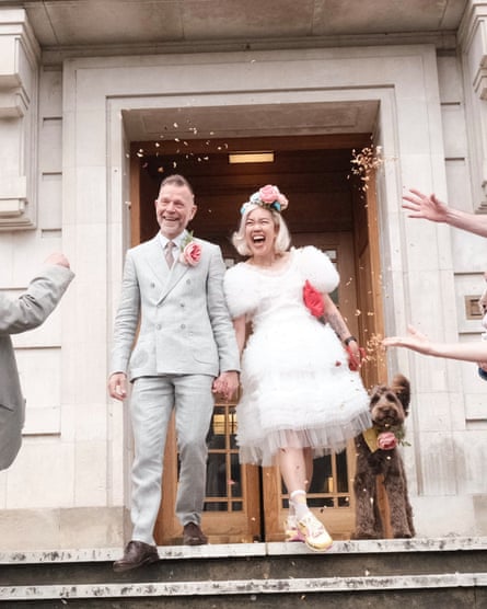 Dominic and Vera in wedding clothes and laughing with their dog on a lead as confetti is thrown over them