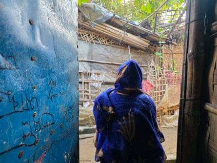 A woman with a blue shawl over her head standing in the doorway of a hut seen from behind